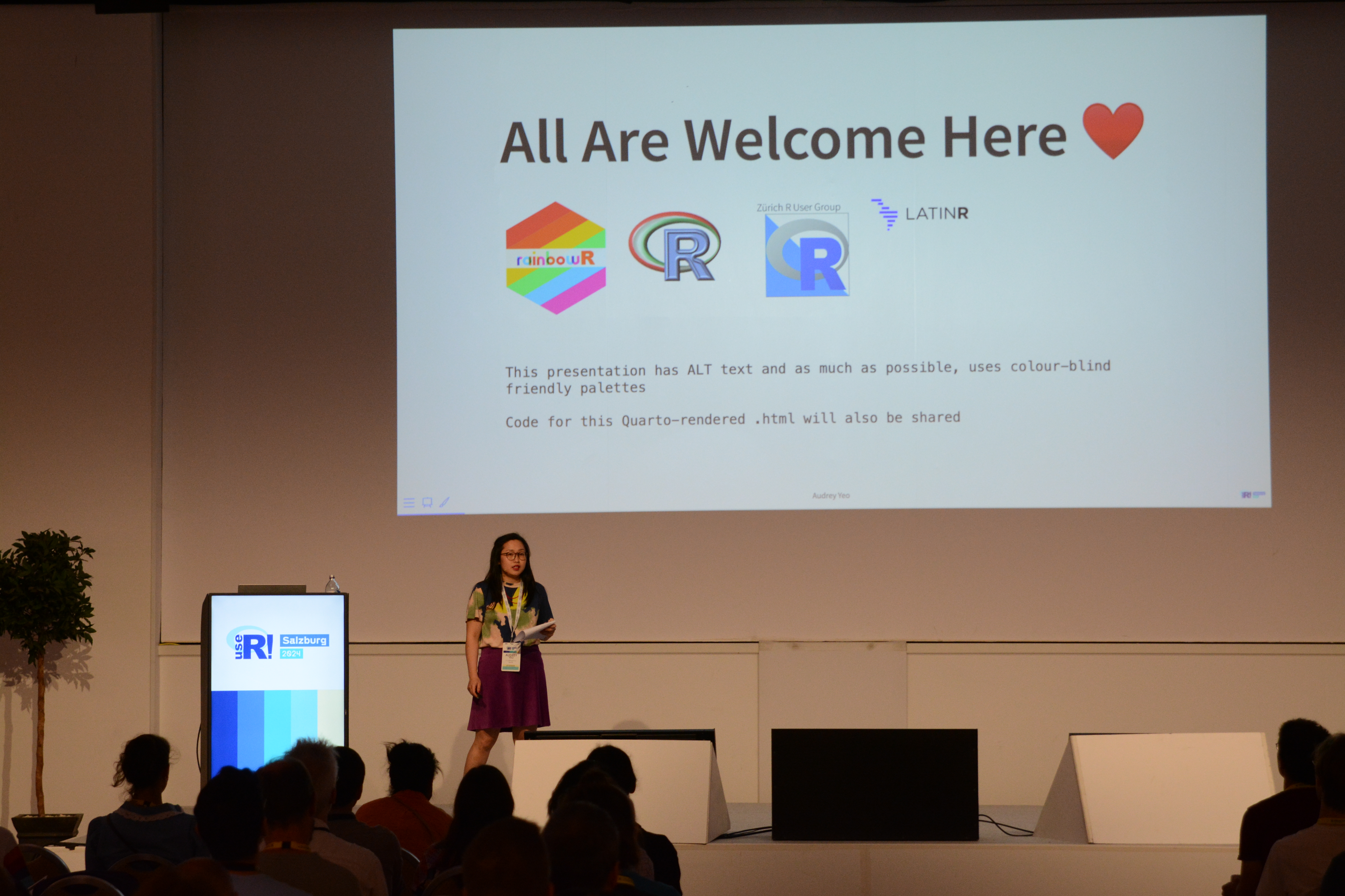 Audrey Yeo on stage at the keynote with slide 'All are Welcome Here'